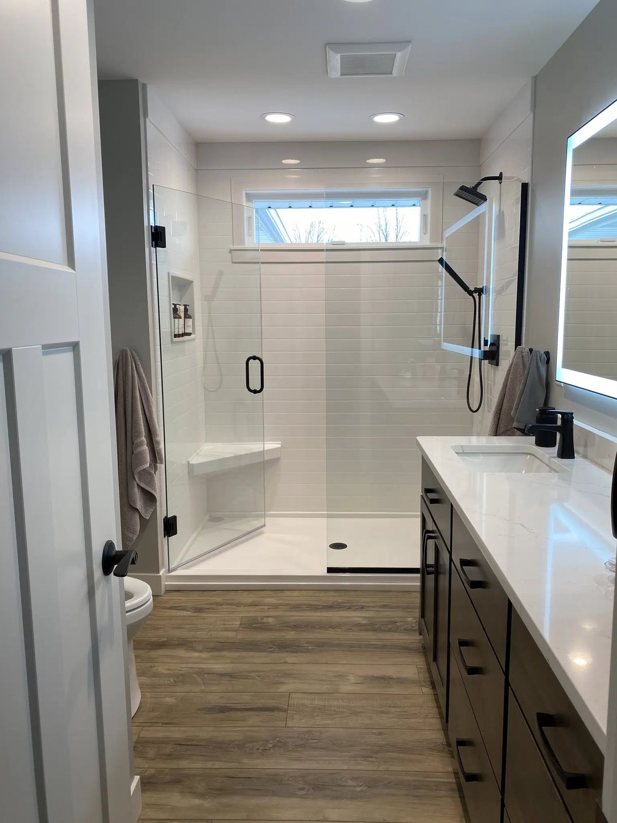 Groutless shower system with white wall panels and a glass enclosure beside a dark wood vanity, available at Sidney Kitchen and Bath in Winchester, VA