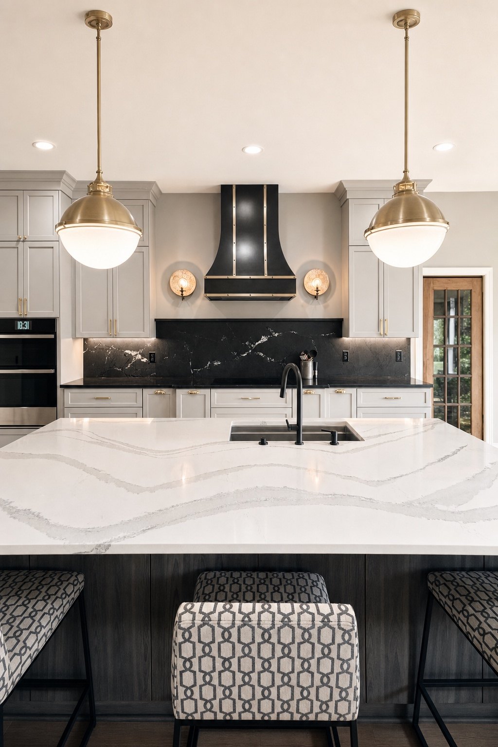 Kitchen island with a veined white countertop under two brass globe pendants, facing a black and brass range hood, by Sidney Design and Build, Winchester, VA